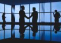 Silhuetas de profissionais cumprimentando-se em um ambiente corporativo com janelas amplas ao fundo, representando networking e acordos em reuniões estratégicas.