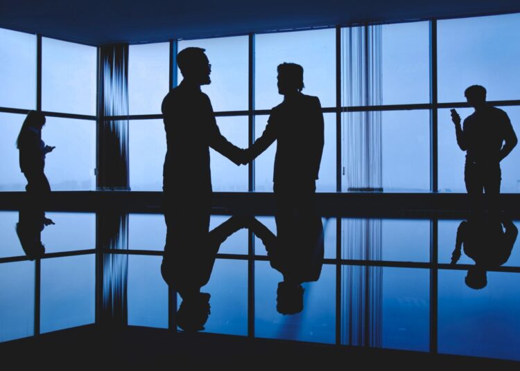 Silhuetas de profissionais cumprimentando-se em um ambiente corporativo com janelas amplas ao fundo, representando networking e acordos em reuniões estratégicas.