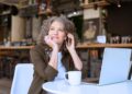 Mulher empreendedora sorridente em café, usando notebook e telefone, representando o protagonismo feminino no empreendedorismo e trabalho remoto.