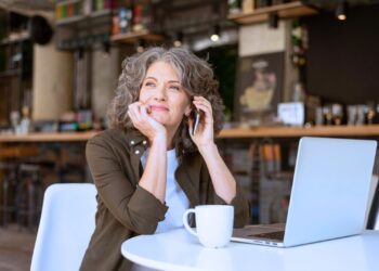 Mulher empreendedora sorridente em café, usando notebook e telefone, representando o protagonismo feminino no empreendedorismo e trabalho remoto.