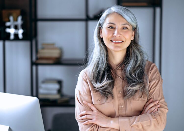Mulher confiante com cabelos grisalhos cruzando os braços, representando a geração X no mercado de trabalho e sua experiência profissional.
