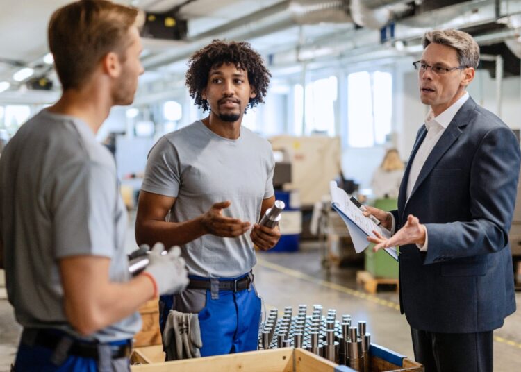 Gestor discutindo com equipe de colaboradores em fábrica sobre processos de gestão de fornecedores e produção industrial.
