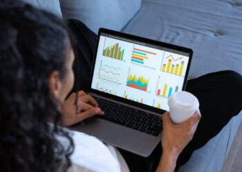 Mulher analisando gráficos financeiros em um laptop enquanto segura um copo de café, representando planejamento financeiro e gestão de investimentos para 2025.