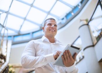 Empresário sorridente segurando um tablet em um ambiente corporativo com teto de vidro, simbolizando inovação e planejamento financeiro.