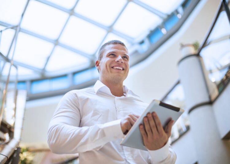 Empresário sorridente segurando um tablet em um ambiente corporativo com teto de vidro, simbolizando inovação e planejamento financeiro.