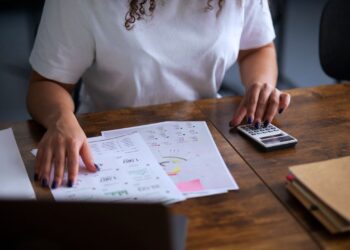 Mulher analisando gráficos financeiros com uma calculadora sobre uma mesa de madeira, representando gestão de capital próprio e capital de terceiros em finanças empresariais.