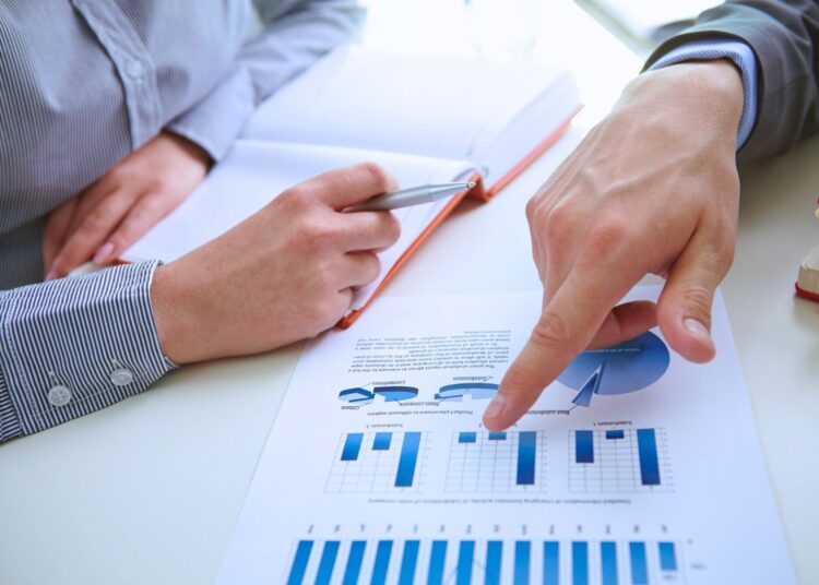 Discussão sobre gráficos financeiros: duas pessoas analisam dados de endividamento geral em um documento com gráficos de barras e setores, destacando tendências financeiras e índices.