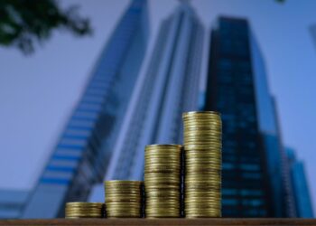 Pilhas crescentes de moedas douradas em primeiro plano, com arranha-céus modernos desfocados ao fundo, simbolizando estrutura de capital, crescimento financeiro e investimentos corporativos.