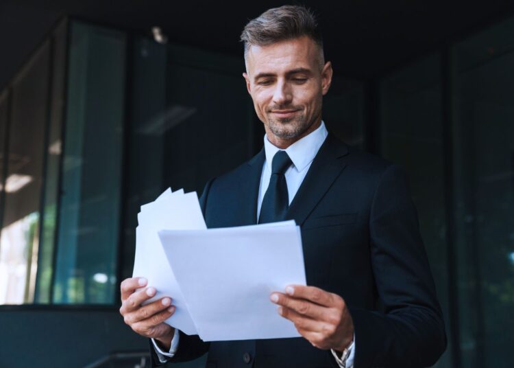 Homem de terno preto segurando documentos e sorrindo, representando análise de capital social e planejamento empresarial.