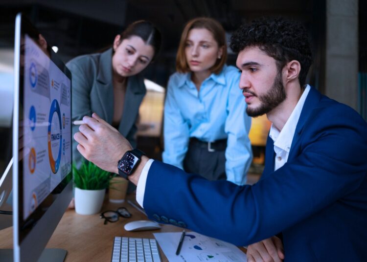 Equipe analisando KPIs de vendas em monitor de computador, discutindo estratégias e performance comercial em ambiente corporativo.