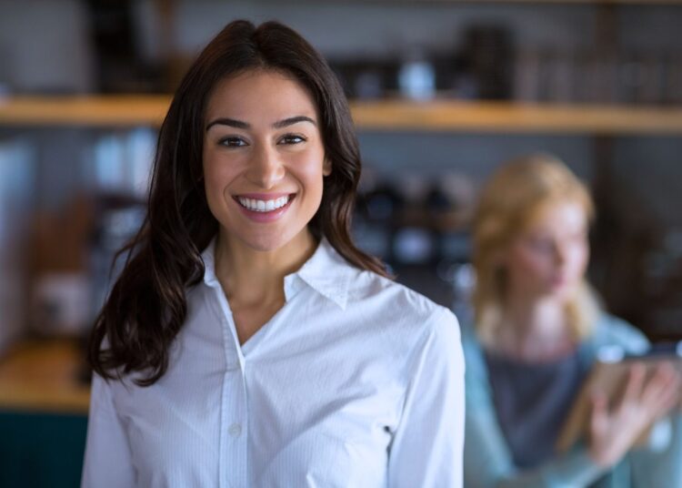 Mulher sorridente vestindo camisa branca em ambiente de trabalho, representando liderança coaching e desenvolvimento profissional.