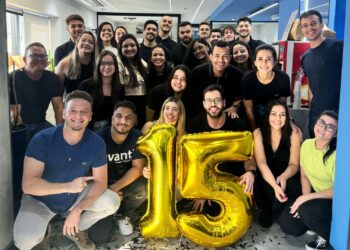 Equipe da Avanti Open Banking reunida em ambiente de escritório para comemoração, com todos sorrindo e segurando balões dourados em forma do número 15, celebrando uma conquista especial.