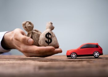 Mão segurando sacos de dinheiro com símbolo de cifrão em direção a um carro de brinquedo vermelho, representando empréstimo com garantia de veículo.