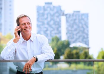 Homem maduro de camisa branca falando ao celular em sacada com prédios ao fundo, representando negociação de compra e venda de imóvel.