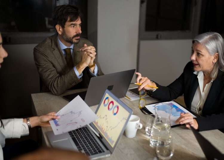 Profissionais em reunião de negócios analisando gráficos financeiros em laptops e papéis, discutindo estrutura de capital e estrutura financeira.