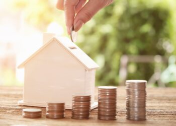 Mão colocando uma moeda em uma casa em miniatura de madeira, com pilhas de moedas ao lado representando crescimento financeiro e permuta de imóvel.