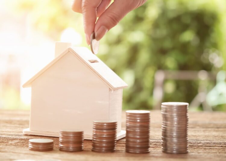 Pessoa inserindo moeda em cofre no formato de casa, ao lado de pilhas crescentes de moedas, simbolizando economia e investimento seguro; imagem representa o diferencial do empréstimo na Avanti com foco em crescimento financeiro e garantia de imóvel.