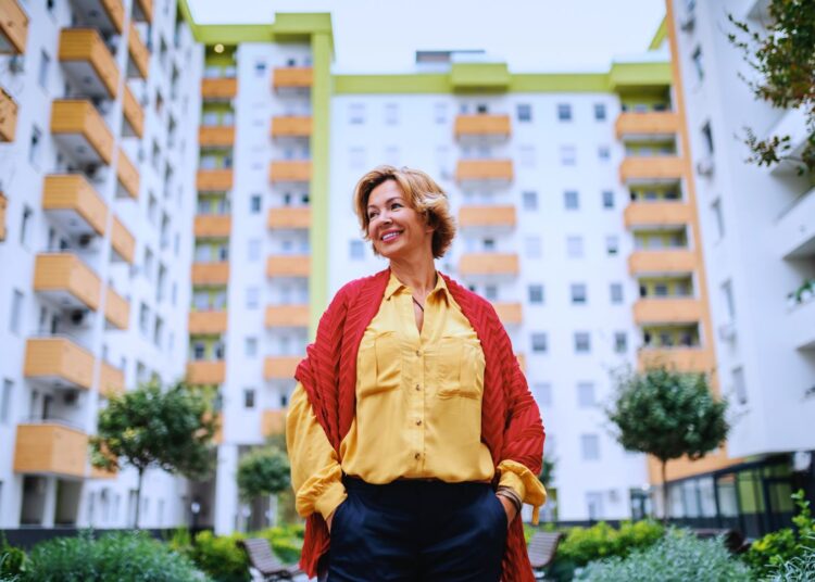 Mulher madura sorridente em frente a prédio residencial moderno, simbolizando área construída.