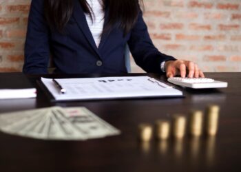 Pessoa em traje formal digitando em uma calculadora sobre uma mesa com documentos, notas de dólar desfocadas à frente e pilhas crescentes de moedas, simbolizando análise de capital de giro.