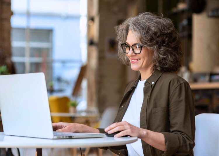 Mulher madura sorridente usando notebook em ambiente moderno e confortável, simbolizando a busca pelo melhor banco para empréstimo com garantia de imóvel.