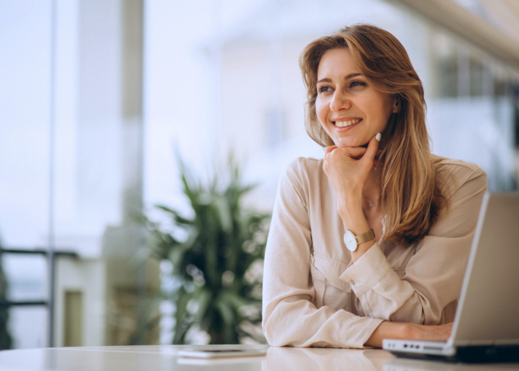 Profissional sorridente em escritório, com notebook aberto, apoiando o queixo com a mão, sugerindo planejamento e estratégia de crescimento com alavancagem financeira,